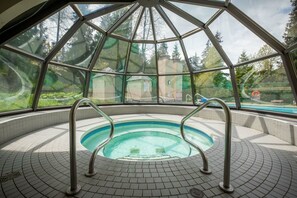 A hot tub with metal railings, surrounded by a tiled floor, under a glass dome with a view of trees and a building. - Mountain views | Pool, hot tub & gym | Ski-in/out (Whistler)