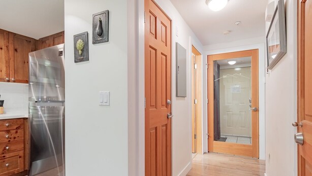 Una cocina con refrigerador de acero inoxidable, armarios de madera y encimera blanca. Se observan cuadros enmarcados en la pared y una puerta de madera que da a otra habitación.