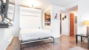 Una habitación compacta con una cama, un escritorio y una kitchenette al fondo.