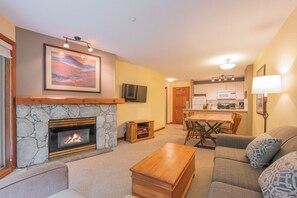 A living room with a stone fireplace, a television, a dining area with chairs, and a kitchen in the background. - Ski-in/out apartment | Shared Hot Tub, Pool & Gym | Superb location (Whistler)