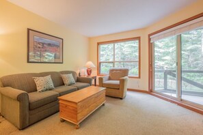 A living room with a sofa, armchair, and coffee table, large windows with a view of trees, and a framed picture on the wall. - Ski-in/out apartment | Shared Hot Tub, Pool & Gym | Superb location (Whistler)
