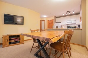 A dining area with a wooden table and chairs, a television mounted on the wall, and a kitchen with white cabinets and appliances. - Ski-in/out apartment | Shared Hot Tub, Pool & Gym | Superb location (Whistler)
