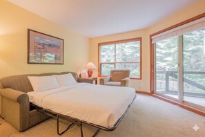 A room with a bed, a chair, a nightstand with a lamp, and a window with a view of trees. - Ski-in/out apartment | Shared Hot Tub, Pool & Gym | Superb location (Whistler)