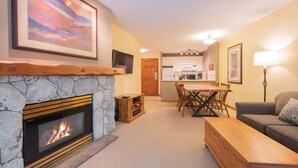 A living room with a stone fireplace, a dining area with a wooden table and chairs, a kitchen with white cabinets, and a grey sofa. - Ski-in/out apartment | Shared Hot Tub, Pool & Gym | Superb location (Whistler)