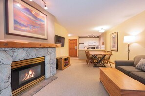 A living room with a stone fireplace, a dining area with a wooden table and chairs, a kitchen with white cabinets, and a grey sofa. - Ski-in/out apartment | Shared Hot Tub, Pool & Gym | Superb location (Whistler)