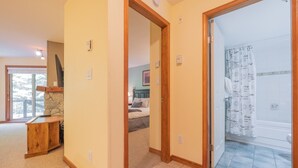 A hallway leading to a bedroom with a stone fireplace, a wooden desk, and a bathroom with a shower curtain. - Ski-in/out apartment | Shared Hot Tub, Pool & Gym | Superb location (Whistler)