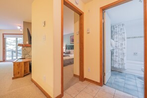 A hallway leading to a bedroom with a stone fireplace, a wooden desk, and a bathroom with a shower curtain. - Ski-in/out apartment | Shared Hot Tub, Pool & Gym | Superb location (Whistler)