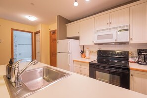 A kitchen with white cabinets, a stainless steel sink, a black stove, and a microwave. - Ski-in/out apartment | Shared Hot Tub, Pool & Gym | Superb location (Whistler)