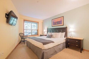 A bedroom with a bed, bedside tables, a chair, a window, and a television mounted on the wall. - Ski-in/out apartment | Shared Hot Tub, Pool & Gym | Superb location (Whistler)