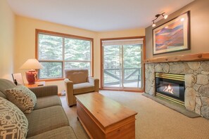 A cozy living room with a stone fireplace, a comfortable sofa, a wooden coffee table, and a large window offering a view of trees. - Ski-in/out apartment | Shared Hot Tub, Pool & Gym | Superb location (Whistler)