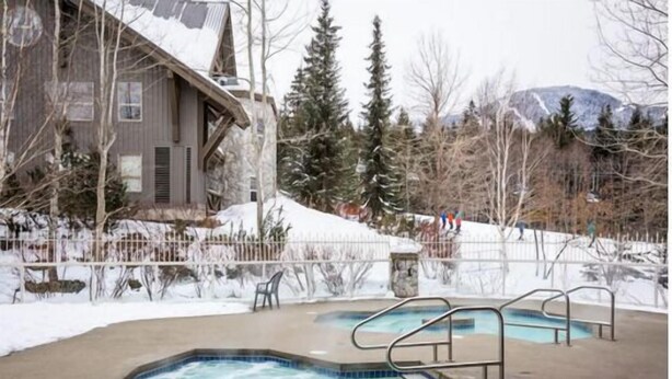 A pool area with two hot tubs, surrounded by a wooden deck and a stone wall. - Ski-in/out apartment | Shared Hot Tub, Pool & Gym | Superb location (Whistler)