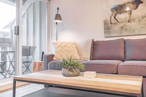 Ein modernes Wohnzimmer mit einer Couch, einem Couchtisch mit Pflanze und Büchern, einer Lampe und einem Bild an der Wand.