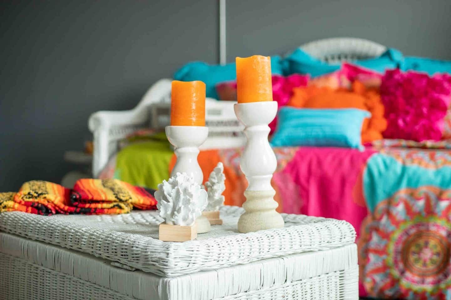 A wicker chair with two white candle holders, a colorful blanket, and a bed with a pink and blue bedspread.