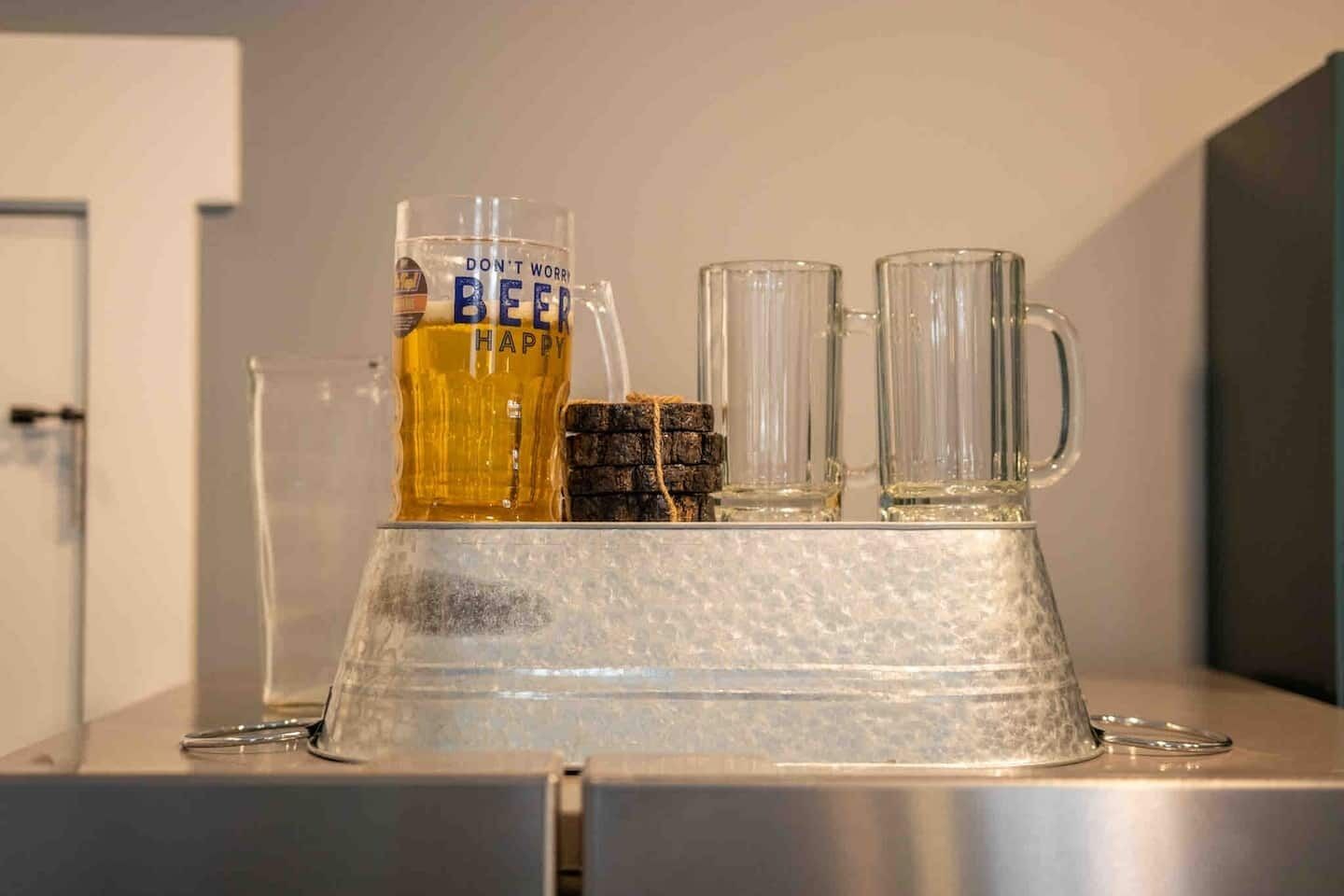 Un espace bar avec un grand verre à bière, deux verres transparents et une bouteille de bière posés sur un comptoir métallique.