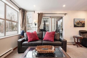 Un salón con un sofá de cuero, dos cojines rojos, una mesa de centro de vidrio, una cesta de mimbre y un cuadro en la pared.