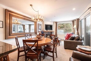 A spacious living room with a fireplace, a dining area with a round table and chairs, and a large window offering a view of the outdoors. - Free parking | Hot tub | Steps to Blackcomb (Whistler)
