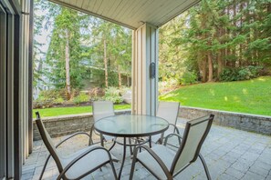 A covered patio with a glass table and chairs, overlooking a lush garden and a forested area. - Steps to Blackcomb | Free Parking | Hot tub (Whistler)