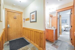 A hallway with wooden doors, a fire extinguisher, a framed picture, and a bathroom with a sink and mirror. - Steps to Blackcomb | Free Parking | Hot tub (Whistler)