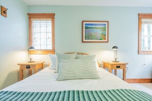 Een slaapkamer met een bed, twee nachtkastjes, een lamp, een foto aan de muur en een raam met jaloezieën.