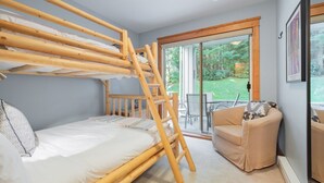Ein Stockbett mit Holzleiter, ein Einzelbett, ein gepolsterter Sessel und eine Glastür zu einem Außenbereich mit Gartenblick.