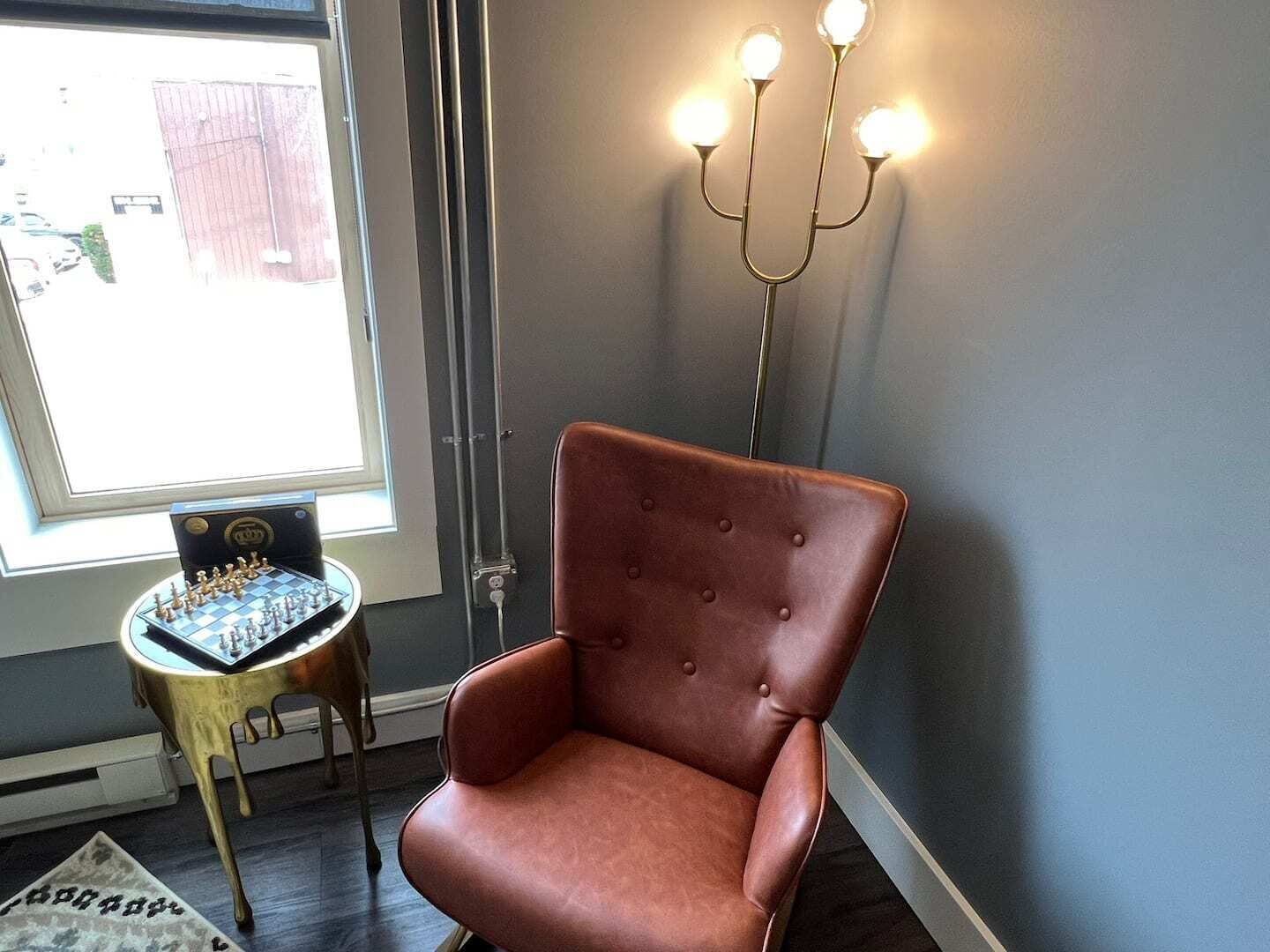 Una habitación con un sillón de cuero marrón, un tablero de ajedrez, una lámpara de pie y una ventana que ofrece vistas a un edificio.