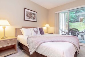 A bedroom with a bed, two bedside lamps, a nightstand, a framed picture on the wall, and a view of greenery through a sliding glass door. - Hot tub | Steps to Blackcomb | Free parking (Whistler)