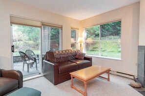 A living room with a leather sofa, a wooden coffee table, and a view of a garden through large windows. - Hot tub | Steps to Blackcomb | Free parking (Whistler)
