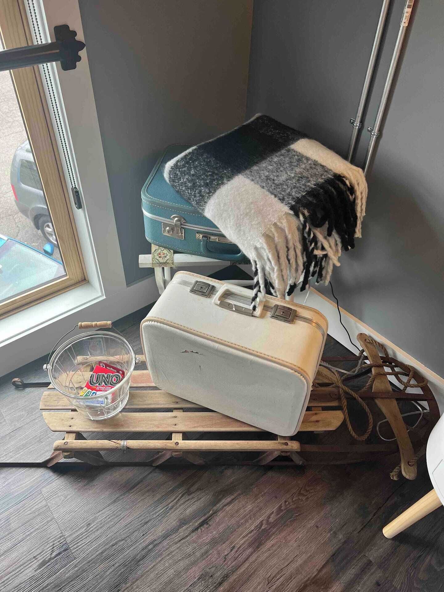 Una stanza con una finestra, una valigia, una coperta, una lampada e un tavolo di legno con una ciotola di vetro.
