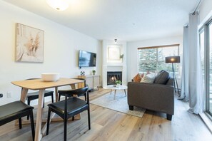 A modern living room with a fireplace, a sofa, a dining area with a table and chairs, and a TV mounted on the wall. - Bright condo | Close to Blackcomb | Free parking (Whistler)
