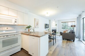 A modern kitchen with a built-in oven, a dishwasher, and a granite countertop. There is a dining area with a table and chairs, and a living room with a sofa and a fireplace. - Bright condo | Close to Blackcomb | Free parking (Whistler)