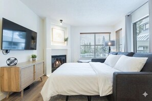 A modern bedroom with a large bed, a flat-screen TV mounted on the wall, a fireplace, and a window with blinds. - Bright condo | Close to Blackcomb | Free parking (Whistler)