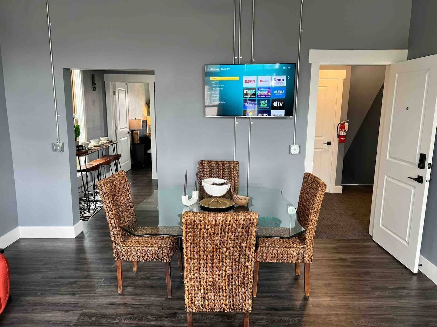 Un comedor moderno con una mesa de vidrio, sillas estampadas y un televisor de pantalla plana montado que muestra varios canales.