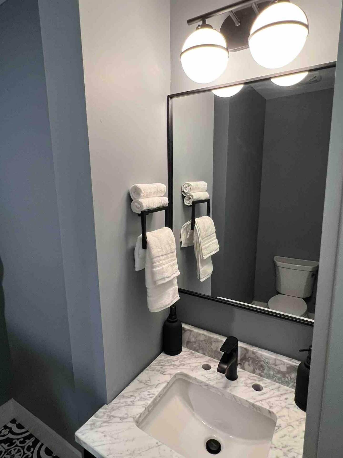 Un baño con lavamanos de mármol, grifo negro y tres toallas blancas colgadas en un perchero negro.
