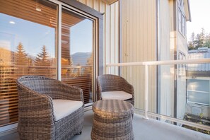 Ein Balkon mit Korbmöbeln und einer Glastür, welche auf Bäume und Berge blickt.