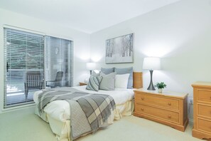A bedroom with a bed, bedside tables, a nightstand, a painting, and a window with blinds. - Stylish | Steps to Blackcomb | Free Parking (Whistler)