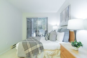 Ein Schlafzimmer mit einem Bett, Nachttischlampen, einem Fenster mit Jalousien, einem Bild an der Wand und einer kleinen Pflanze auf dem Nachttisch.