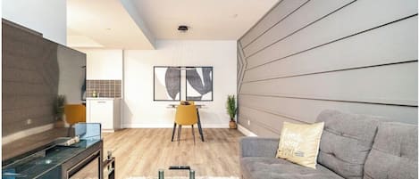 A modern living room with a grey sofa, a coffee table, a TV, and a dining area with a yellow chair.