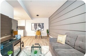 A modern living room with a grey sofa, a coffee table, a TV, and a dining area with a yellow chair. - Cozy & Stylish 1-Bedroom Suite Perfect for Couples or Solo Travellers (Calgary)