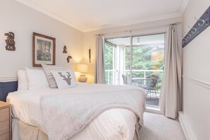 A bedroom with a bed, a nightstand, a lamp, a painting, and a view of the outdoors through a sliding glass door. - Modern 2BDR + Loft | Close to Ski Lifts and Shops (Whistler)