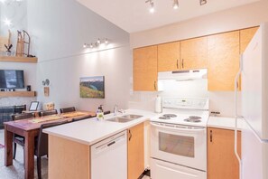 Una cocina moderna con electrodomésticos blancos, armarios de madera, un comedor con mesa y sillas, y un televisor de pantalla plana montado en la pared.