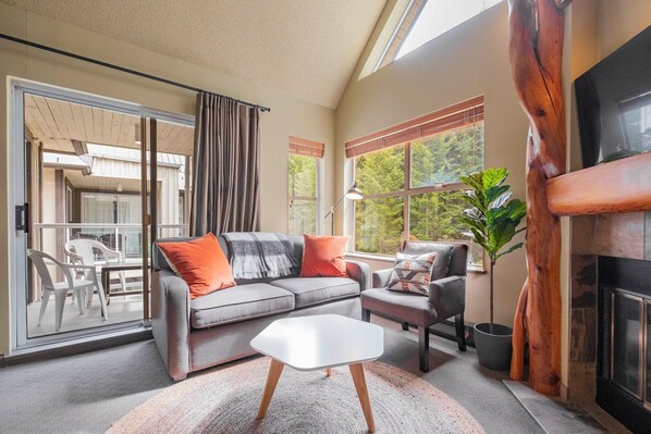 A living room with a sofa, armchair, and a small table. There is a sliding glass door leading to an outdoor area with chairs. A fireplace is visible on the right side. - Complex hot tub | Close to Lifts | Free Parking (Whistler)