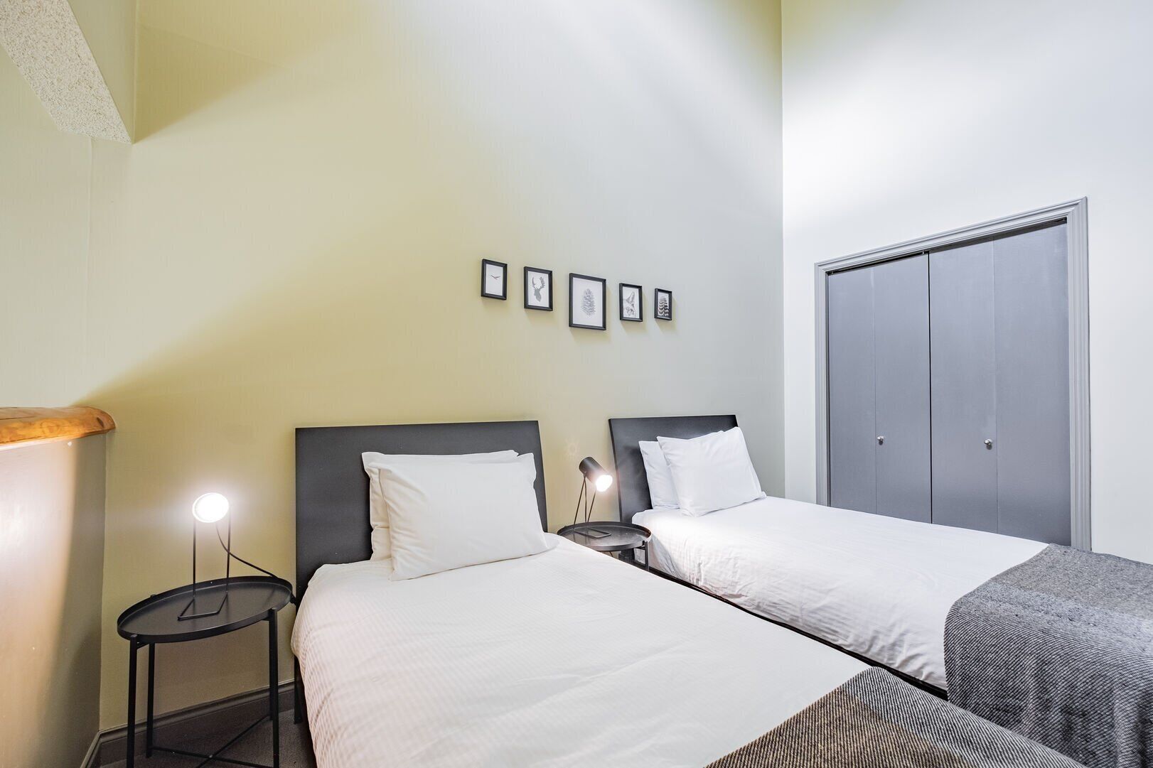 A hotel room with two beds, a small table with a lamp, a wardrobe, and framed pictures on the wall.