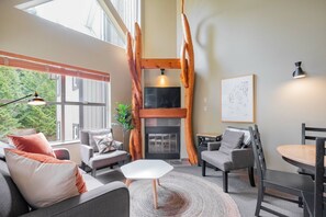 A modern living room with a fireplace, a dining area, and a large window with a view of trees. - Complex hot tub | Close to Lifts | Free Parking (Whistler)