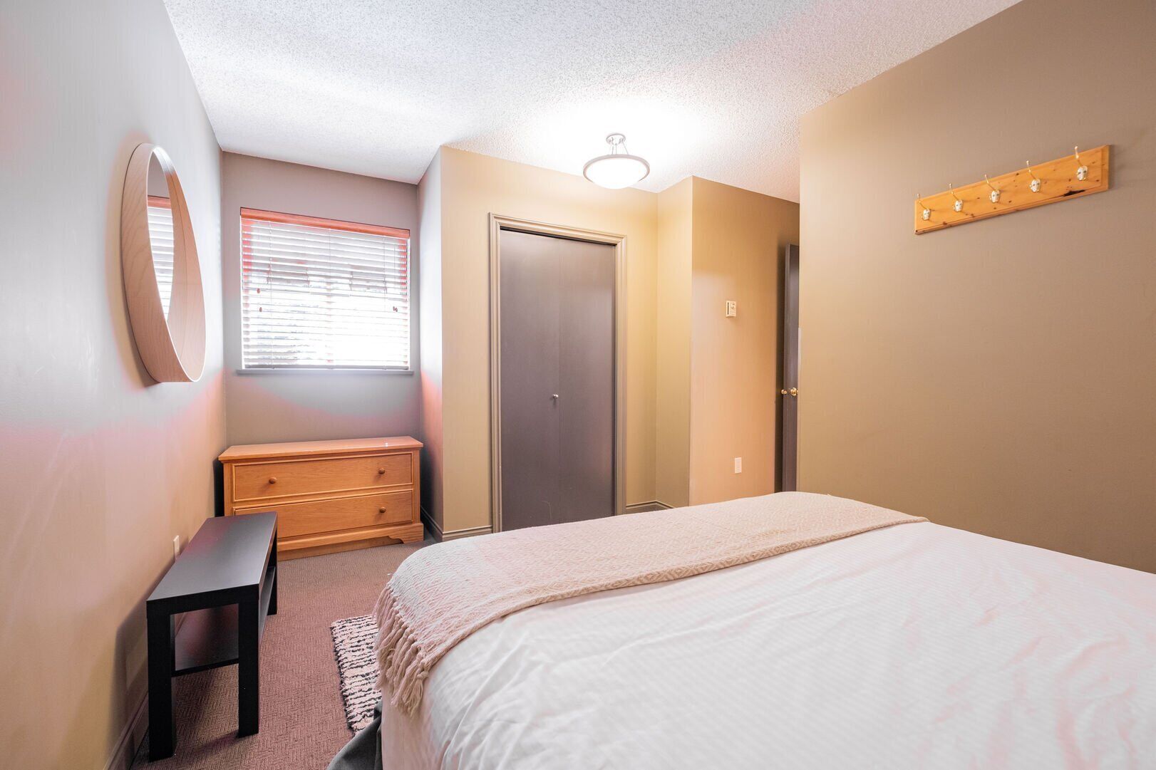 A bedroom with a bed, a dresser, a mirror, a window with blinds, a door, and a light fixture.