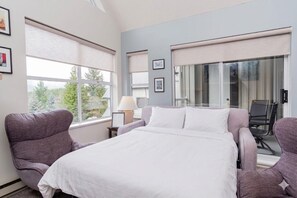 Habitación de hotel con cama, dos sillones, un escritorio con lámpara y ventanas amplias con persianas.