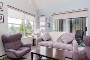 A modern living room with a sofa, armchair, and coffee table, large windows with blinds, and a view of greenery.