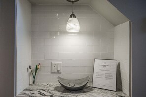 Un baño con encimera de mármol, lavamanos sobre encimera, lámpara y un cartel de bienvenida.