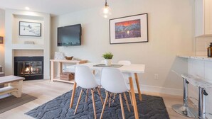 A modern living room with a fireplace, a dining area with white chairs, a TV mounted on the wall, and a framed painting above the fireplace. - Newly renovated 1 bedroom near Blackcomb Gondola - less than 5 min walk (Whistler)