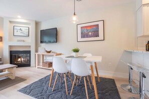 Una sala de estar moderna con chimenea, un comedor con sillas blancas, un televisor montado en la pared y un cuadro enmarcado sobre la chimenea.