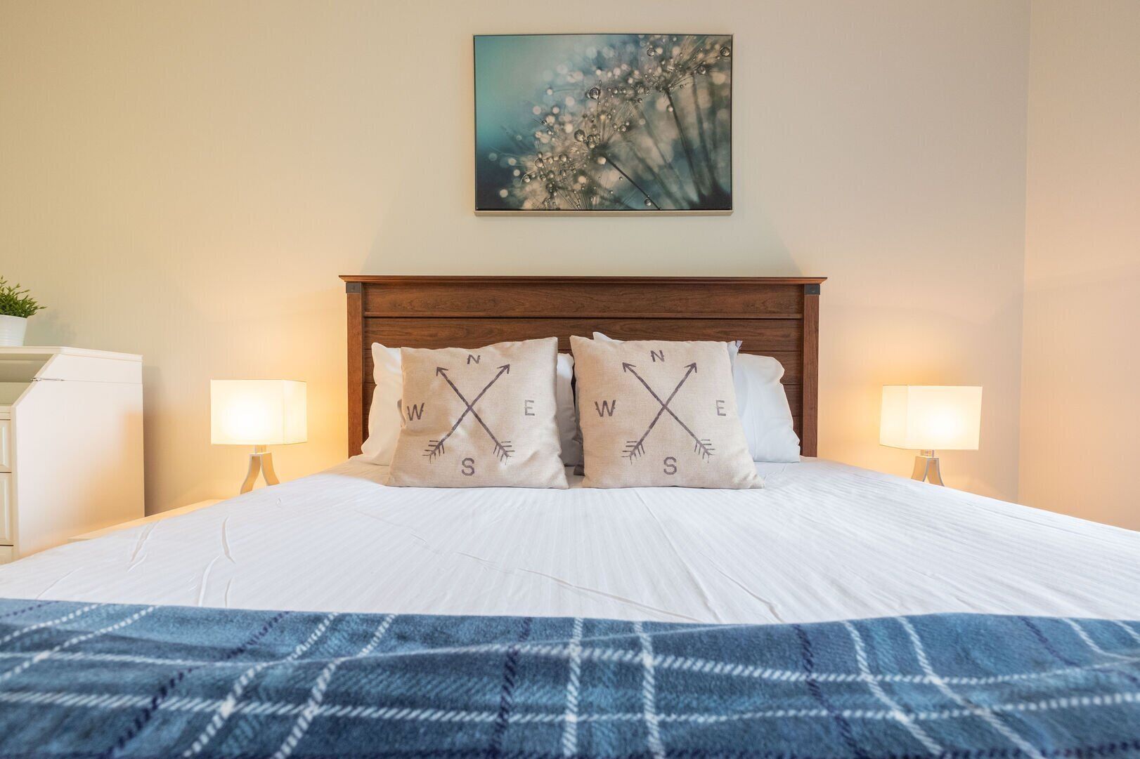 Een net opgemaakt bed met een blauwe deken, twee kussens met kompasrichtingen, een houten hoofdbord, een aan de muur hangend schilderij en bedlampjes.